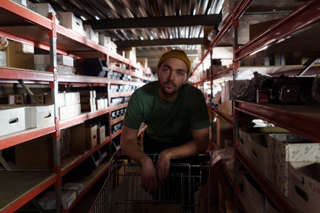 Man in green shirt and beanie pushing a cart in a busy warehouse aisle.